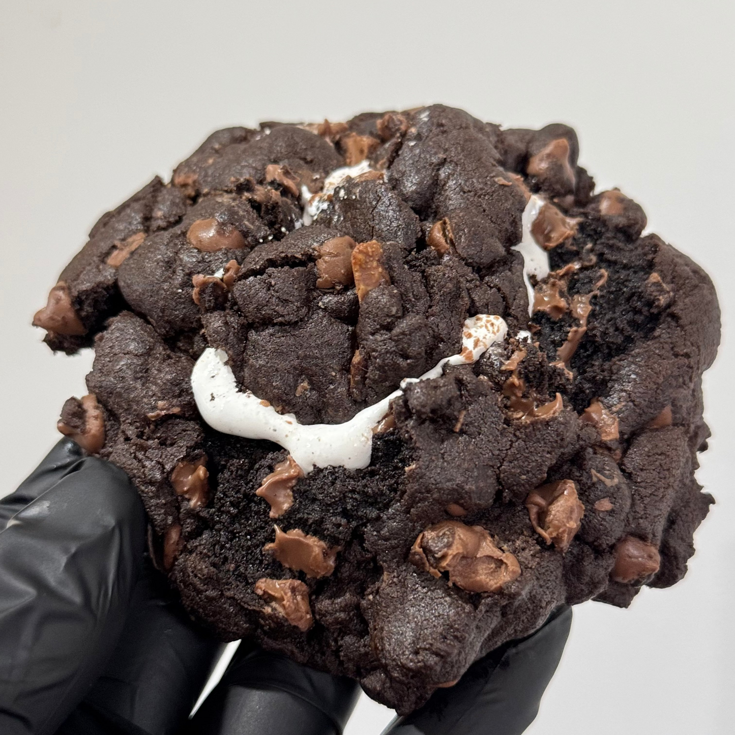 A close-up of a large chocolate cookie with chocolate chips and a white marshmallow center, held by a hand wearing a black glove against a plain light gray background.