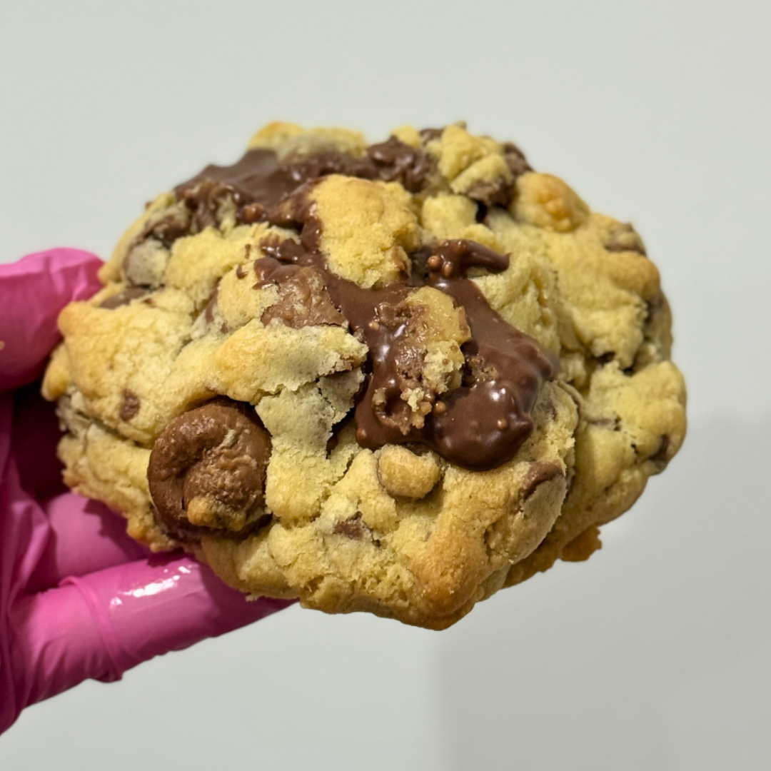 Chocolate chip cookie with a gooey center held by a pink gloved hand against a plain background