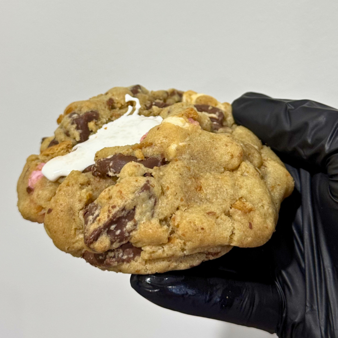 Hand holding a large cookie with chocolate chips and marshmallows, wearing a black glove on a light gray background.