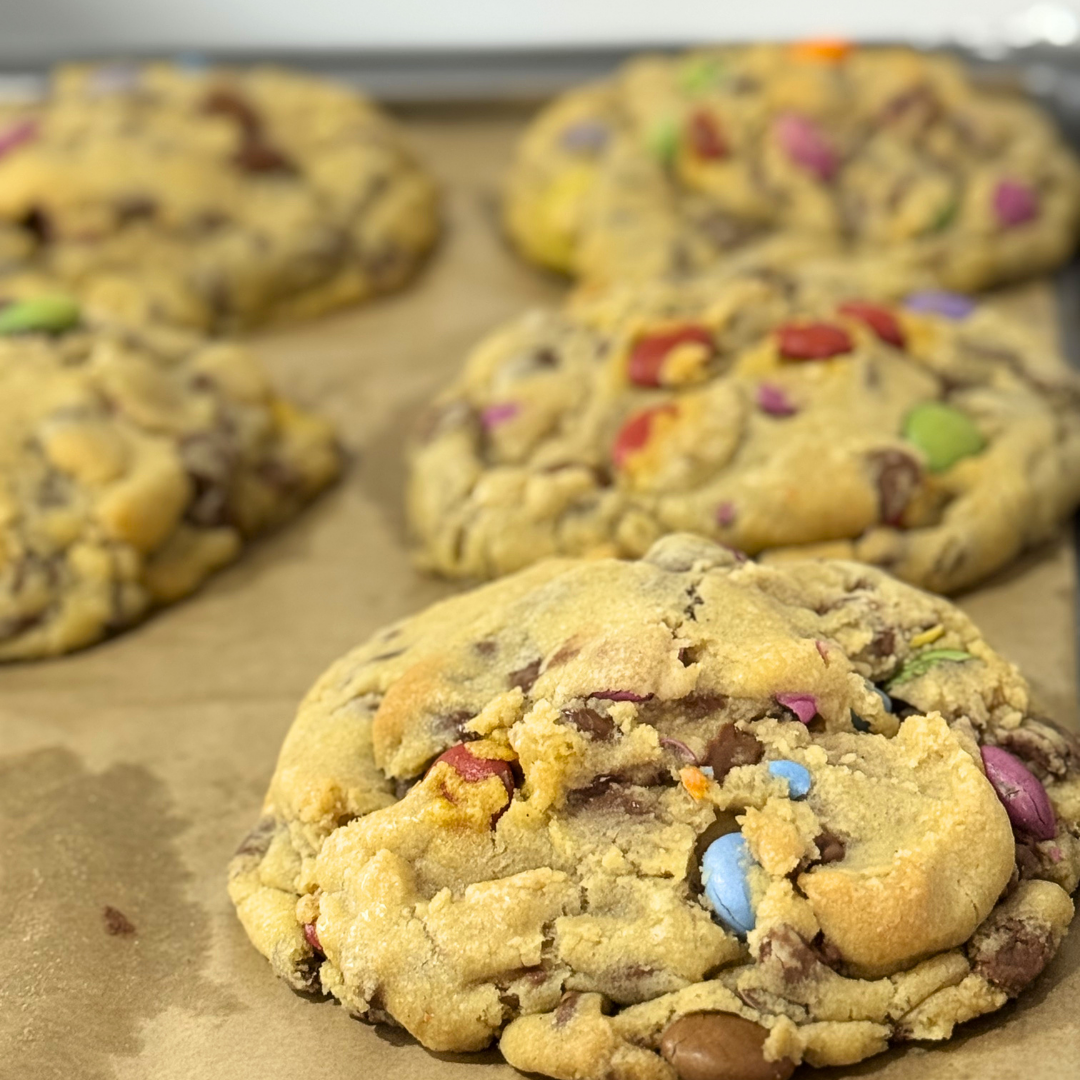 Close-up of several large chocolate chip cookies with colorful M&M's candies on a baking sheet lined with parchment paper.