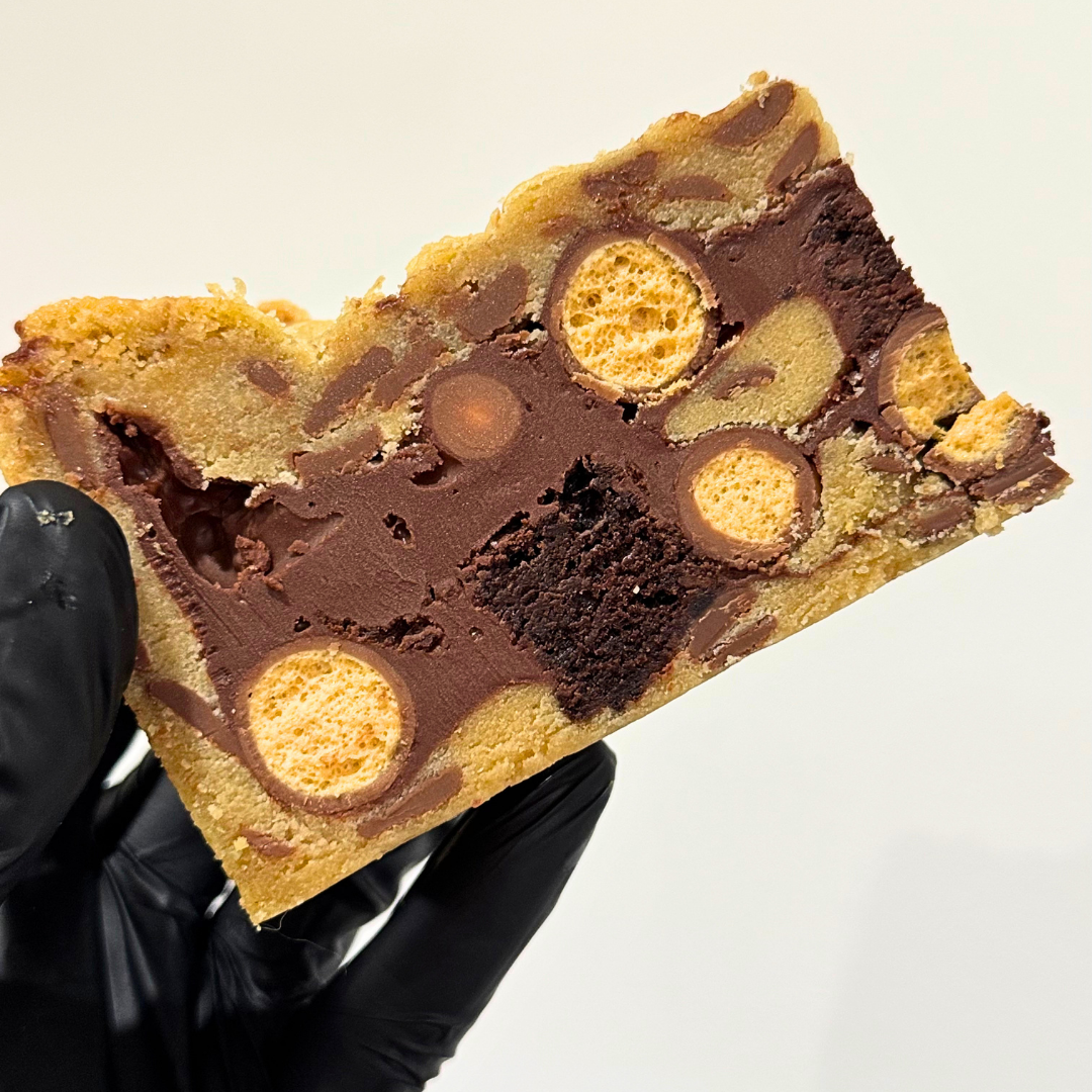 A close-up of a hand wearing a black glove holding a large, square-cut chocolate chip cookie with swirls of dark and milk chocolate and embedded chocolate-coated candy pieces, against a plain white background.