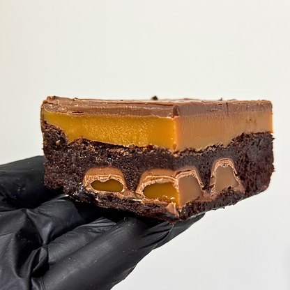 A close-up of a chocolate dessert slice held by a black gloved hand against a plain light background. The dessert has a dark brown chocolate base, a layer of caramel-like amber filling, and a smooth chocolate topping.