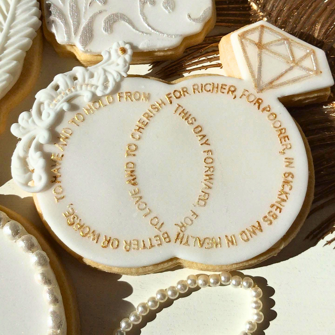 A close-up of an iced cookie shaped like a ring with gold lettering that reads, 'TO LOVE AND TO HOLD FROM THIS DAY FORWARD.