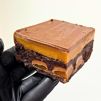 A square chocolate dessert bar with a glossy brown chocolate topping, a thick caramel layer, and a dark chocolate base, held by a hand wearing a black glove against a plain light background.