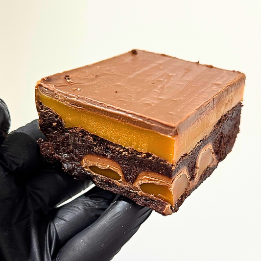 A square chocolate dessert bar with a glossy brown chocolate topping, a thick caramel layer, and a dark chocolate base, held by a hand wearing a black glove against a plain light background.