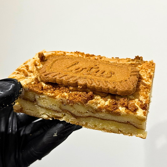 A hand wearing a black glove holds a square slice of a layered dessert cake topped with a biscuit, against a plain light background.
