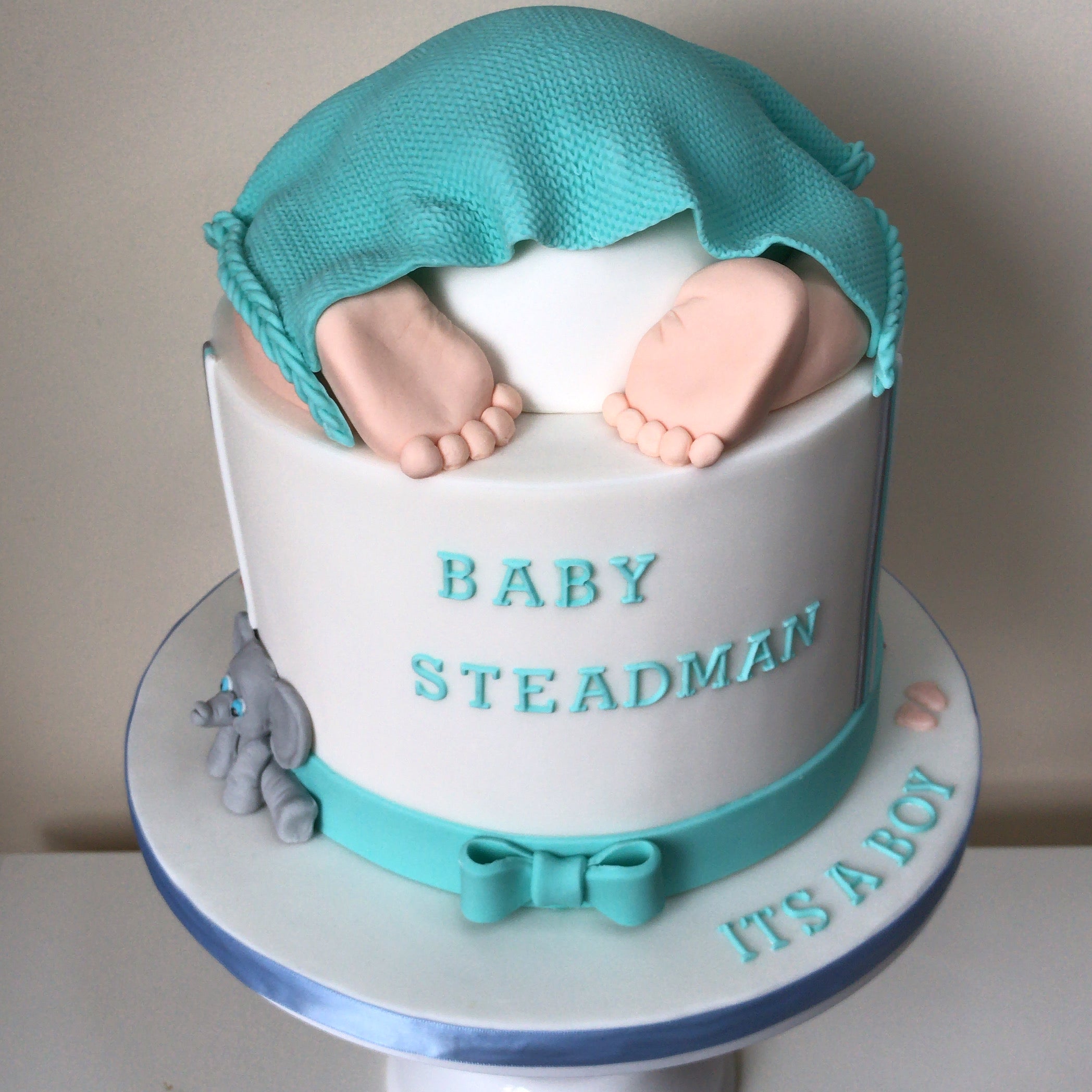 A baby shower cake decorated with a fondant baby's bottom and feet, wrapped in a blue fondant blanket, with 'BABY STEADMAN' and 'IT'S A BOY' written on the side.
