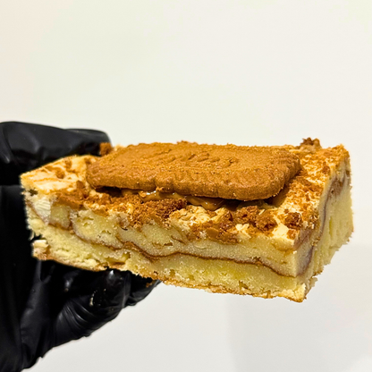 A hand wearing a black glove holds a square slice of layered cake or pastry topped with a biscuit, against a plain light background.