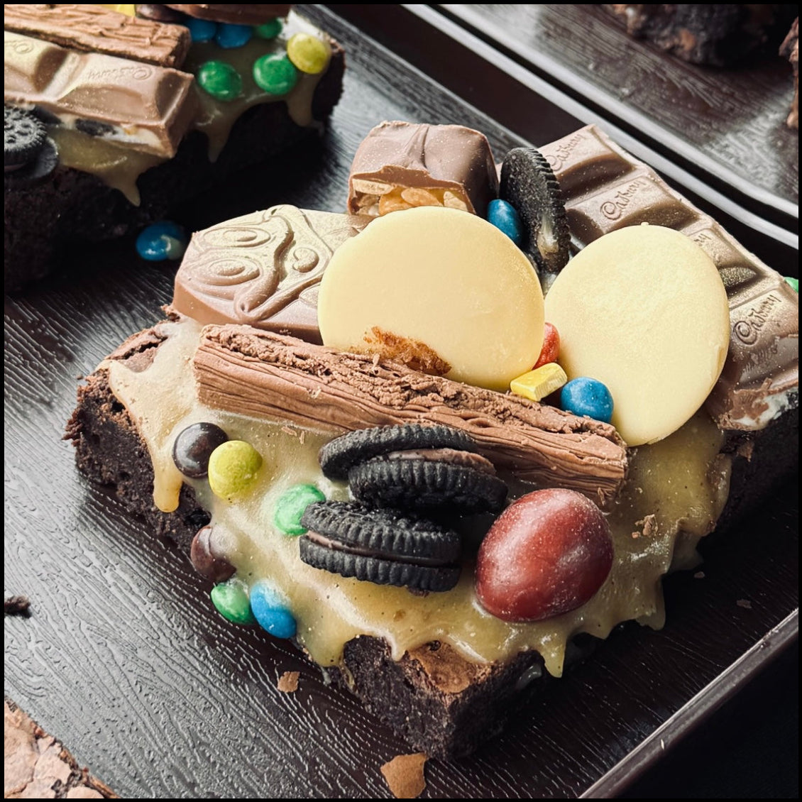 A close-up of a chocolate brownie topped with milk chocolate chunks, white chocolate discs, Oreo cookies, blue candies, and a chocolate stick, served on a dark tray.