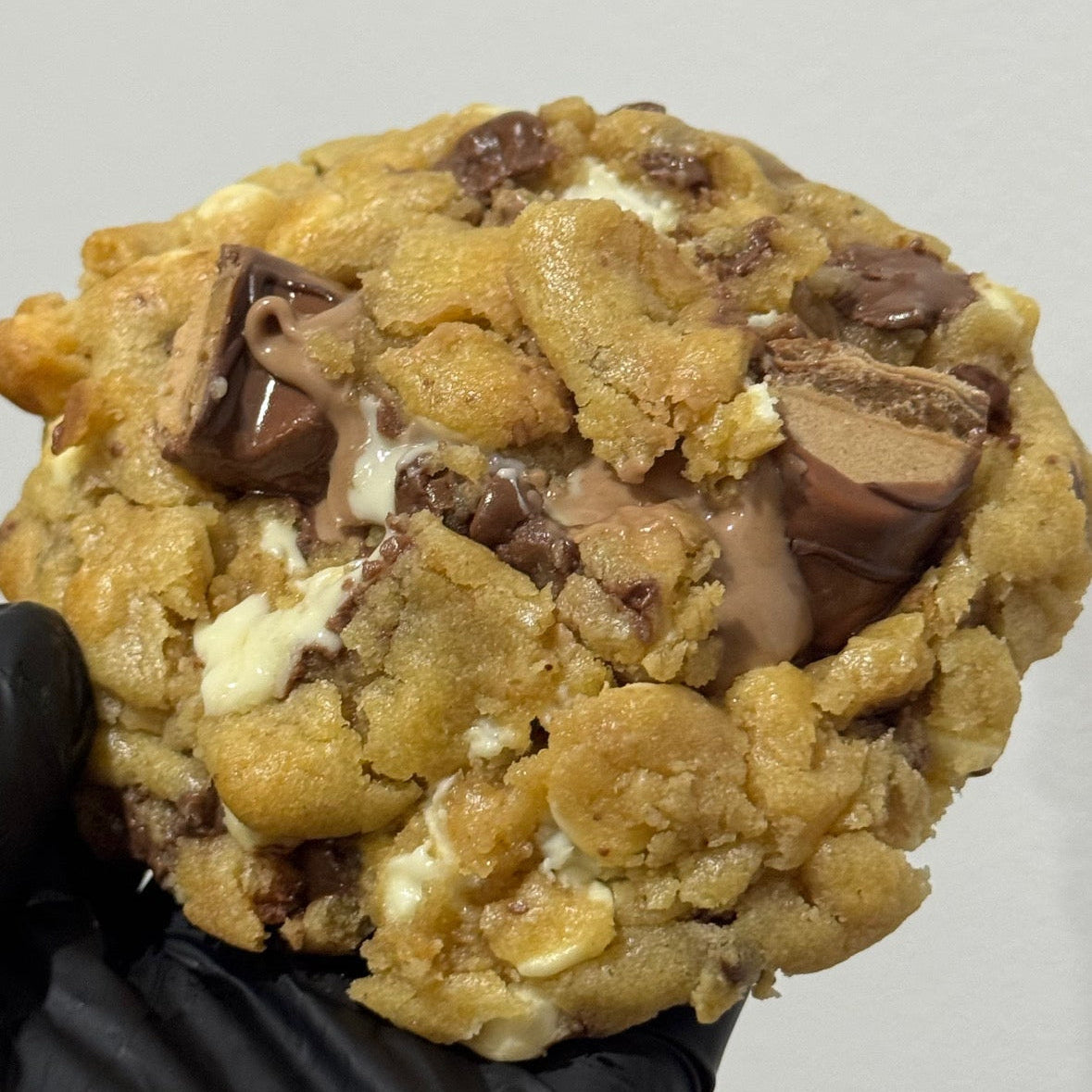 A close-up of a large chocolate chip cookie filled with milk chocolate and white chocolate pieces, held by a hand wearing a black glove against a plain light gray background.