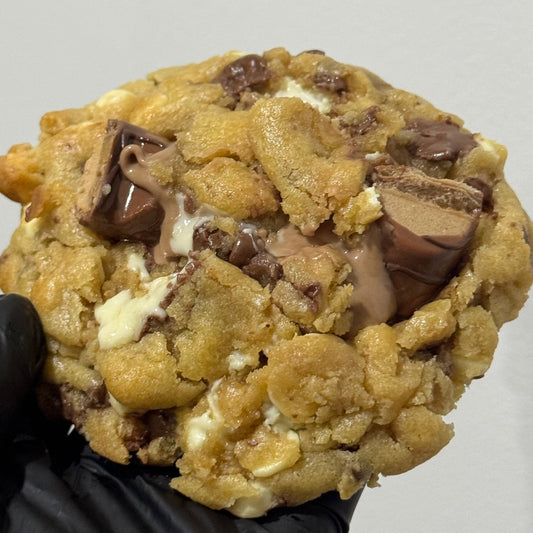 A close-up of a large chocolate chip cookie filled with milk chocolate and white chocolate pieces, held by a hand wearing a black glove against a plain light gray background.
