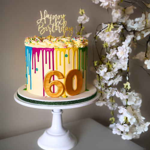 A tall 60th birthday cake with white frosting, colorful rainbow drips, a gold 'Happy Birthday' topper, and large gold '60' numbers, placed on a white cake stand next to white flowers.