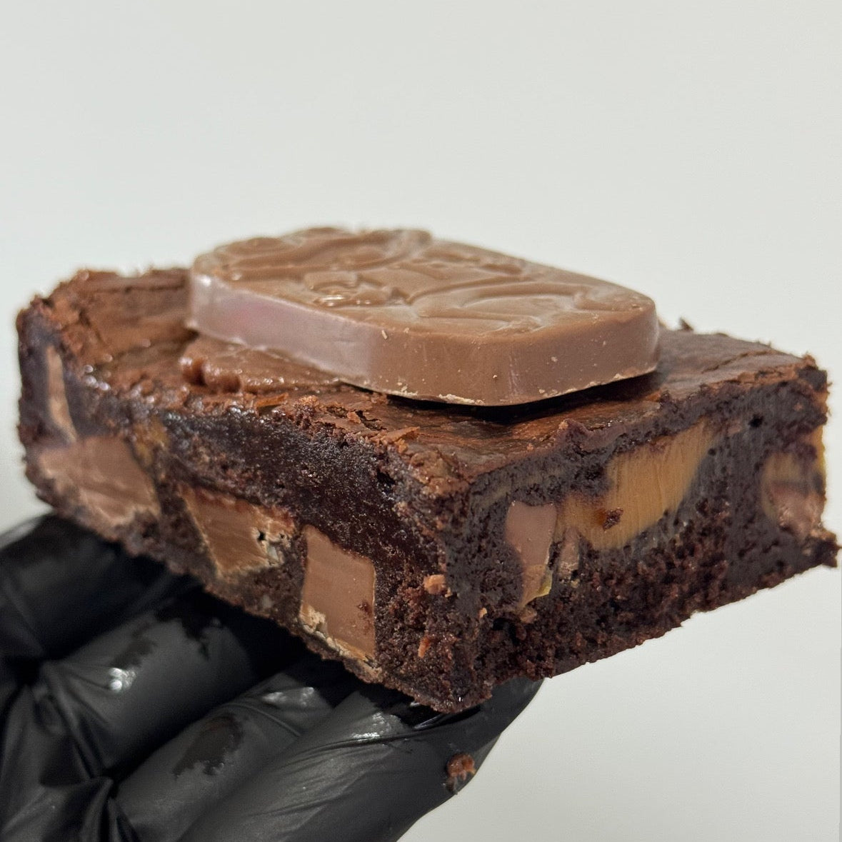 A chocolate brownie topped with a milk chocolate bar, held by a hand wearing a black glove against a plain light gray background.