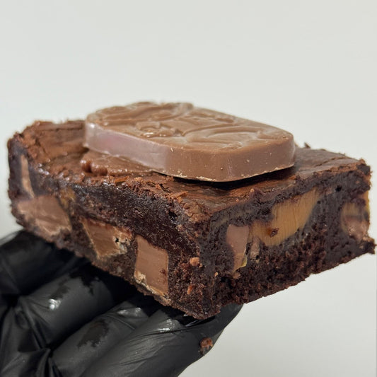 A chocolate brownie topped with a milk chocolate bar, held by a hand wearing a black glove against a plain light gray background.