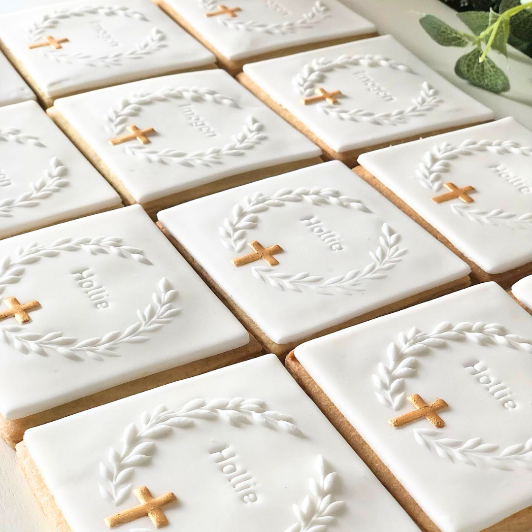 A close-up of multiple rectangular sugar cookies decorated with white icing, featuring a white laurel wreath, a small gold cross, and the name 'Hollie' in script.