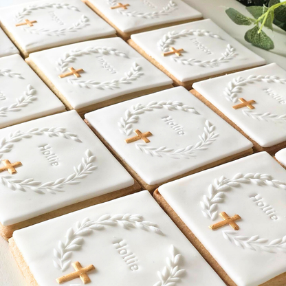 A close-up of multiple rectangular sugar cookies decorated with white icing, featuring a white laurel wreath, a small gold cross, and the name 'Hollie' in script.