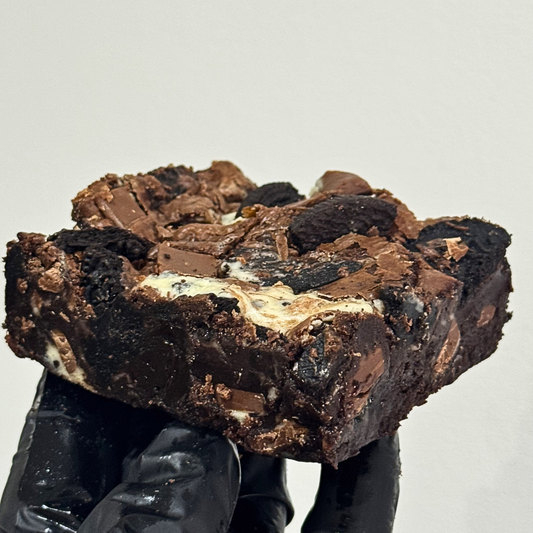 A close-up of a hand in a black glove holding a large, rich brownie topped with chocolate chunks and white cream, set against a plain light gray background.