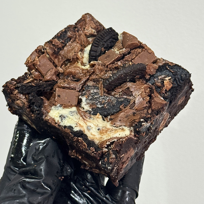 A close-up of a hand in a black glove holding a large square brownie topped with chocolate chunks, cookie crumbs, and a cream-colored filling, against a plain light background.
