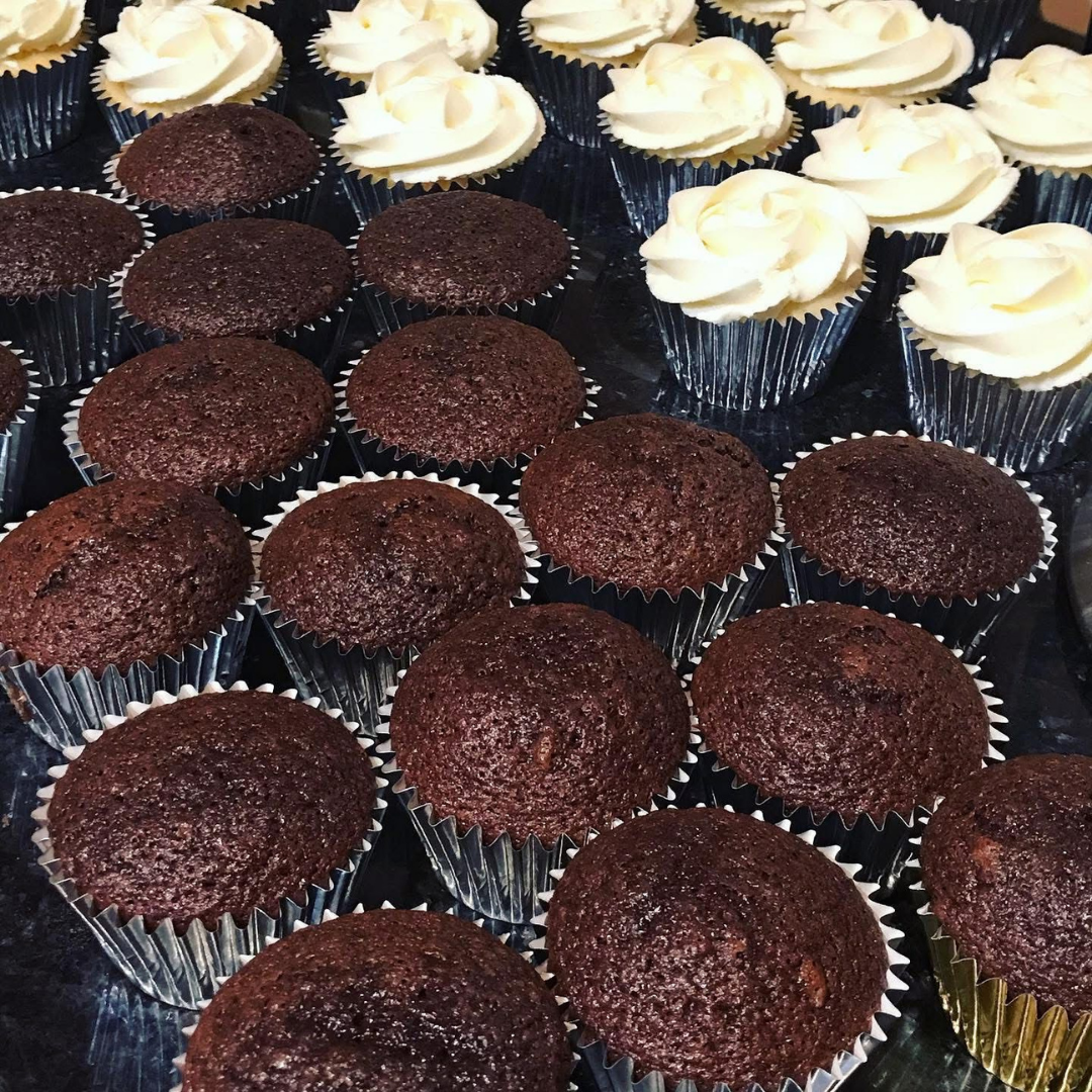 A close-up arrangement of chocolate cupcakes, with several topped in the back row by thick white frosting and others left plain, all in silver and gold paper liners.