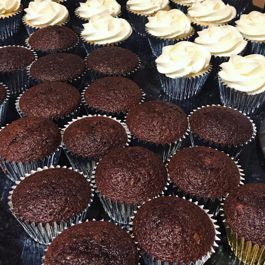 A close-up arrangement of chocolate cupcakes, with several topped in the back row by thick white frosting and others left plain, all in silver and gold paper liners.