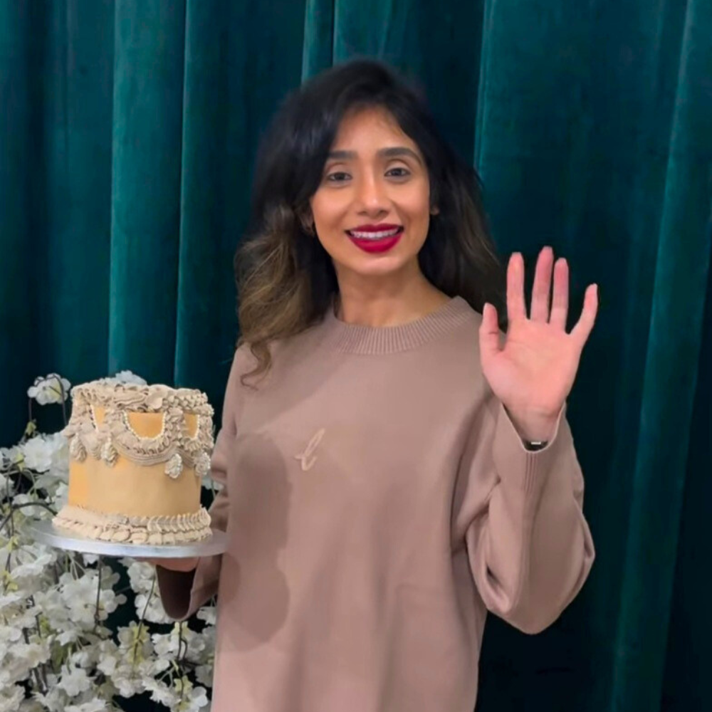 Woman holding a decorated cake and waving in front of a teal curtain