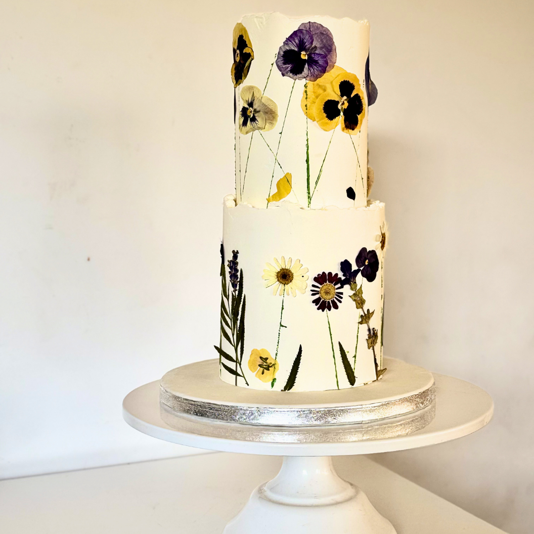 A two-tier white cake decorated with realistic pressed flowers in yellow, purple, and black, displayed on a white cake stand against a plain white background.