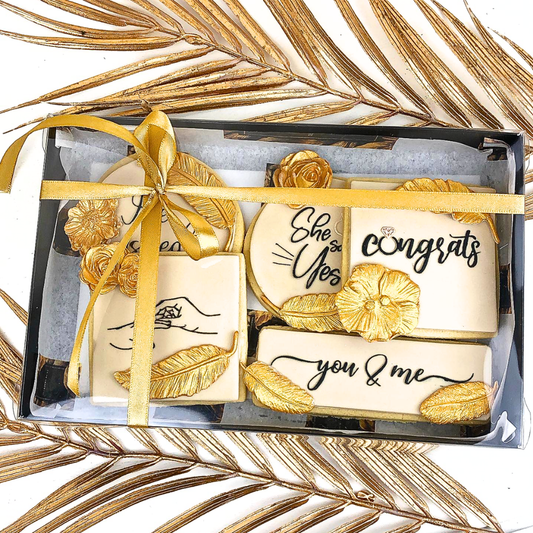 A boxed set of personalized wedding cookies decorated with gold leaf patterns and a gold ribbon, displayed on a white background with gold palm leaves.