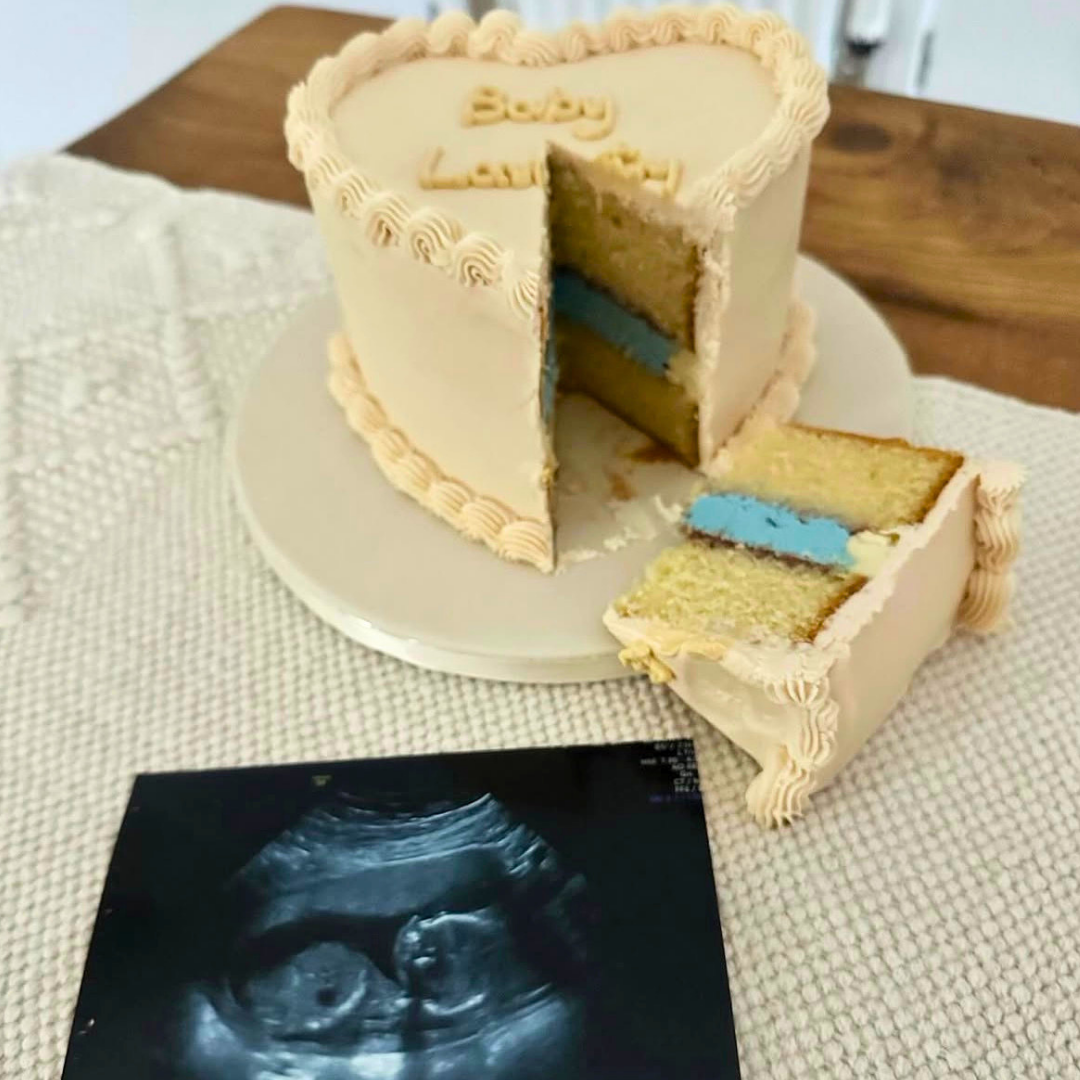 A heart-shaped birthday cake with beige frosting and blue cake layers, with a slice cut out and placed beside it. The top of the cake is written 'Baby Love!'.