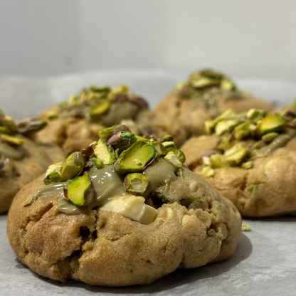 A close-up of a golden-brown cookie topped with chopped green pistachios and a drizzle of glaze, with additional cookies blurred in the background on a light surface.