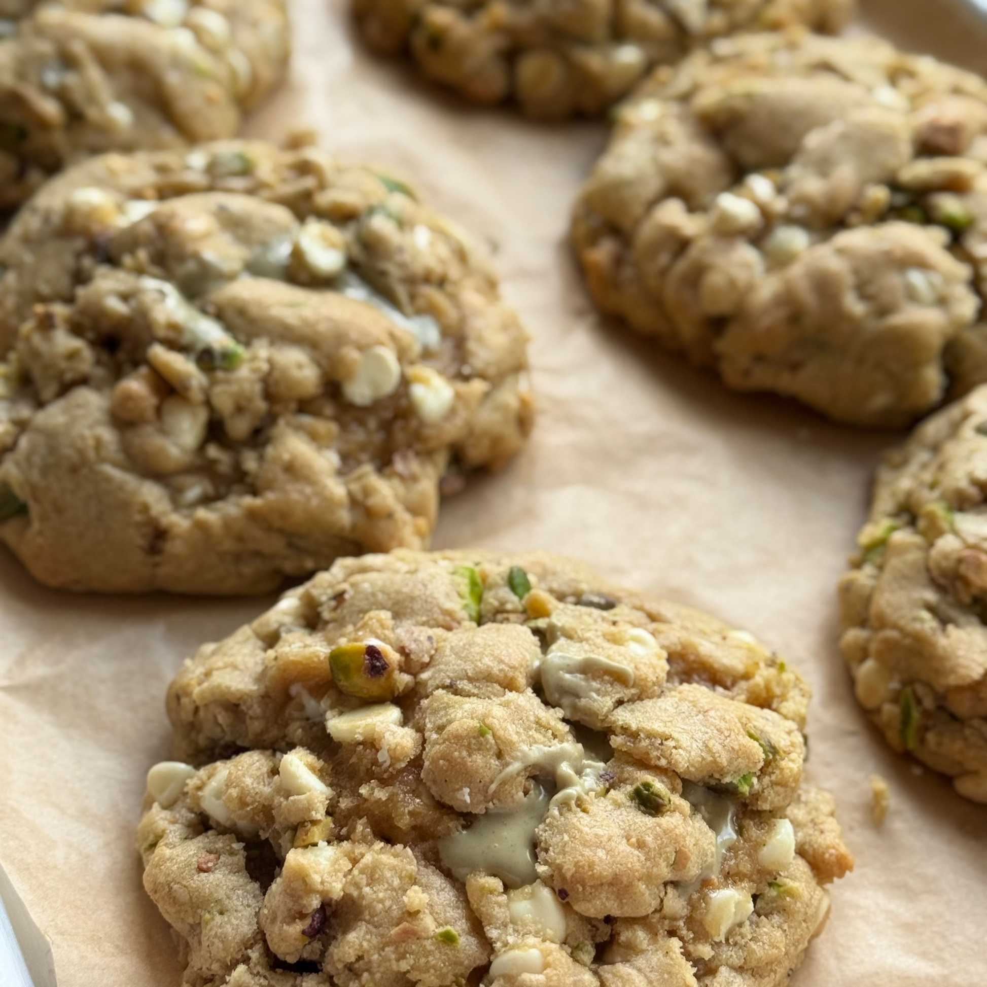 Close-up of several thick, chewy white chocolate chip cookies with nuts and green bits on parchment paper.