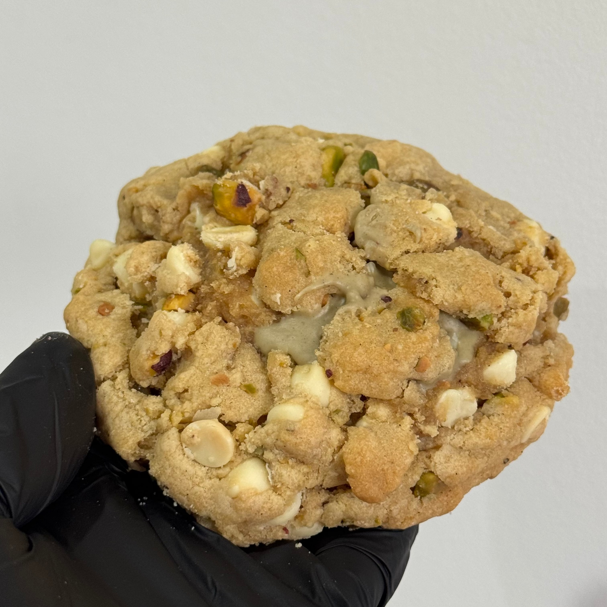 A large, thick white chocolate cookie with nuts and dried fruit pieces, held by a hand wearing a black glove against a plain light gray background.