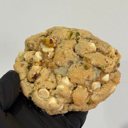 A large, thick white chocolate cookie with nuts and dried fruit pieces, held by a hand wearing a black glove against a plain light gray background.