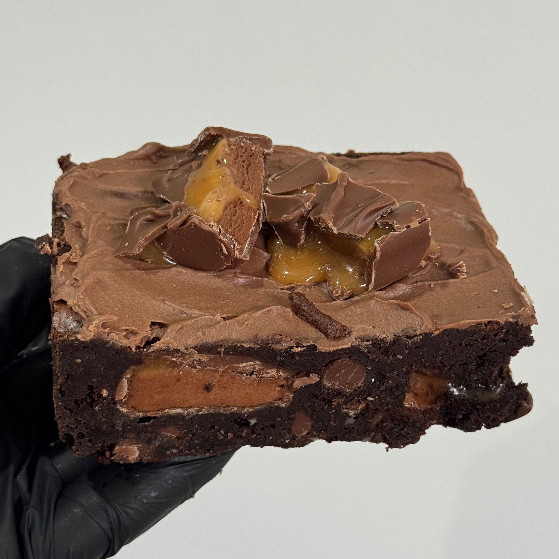 A hand wearing a black glove holds a square chocolate brownie topped with melted chocolate, caramel, and chocolate chunks against a plain light gray background.