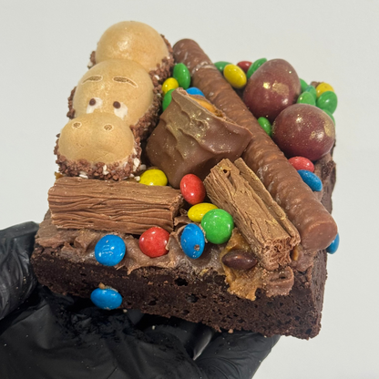 A chocolate brownie topped with assorted candy and chocolate pieces, held by a black gloved hand against a plain light gray background.