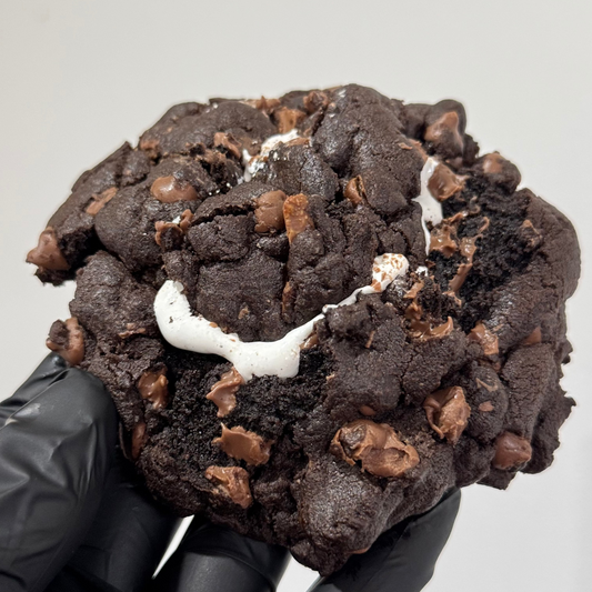 A close-up of a large chocolate cookie with chocolate chips and a white marshmallow center, held by a hand wearing a black glove against a plain light gray background.