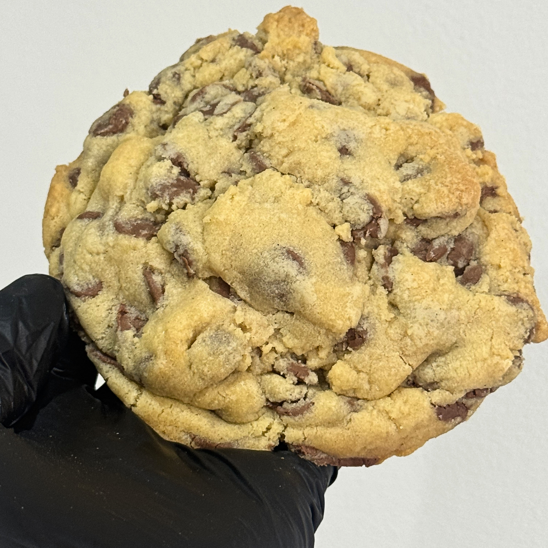 A large chocolate chip cookie held up by a black gloved hand against a plain light background.