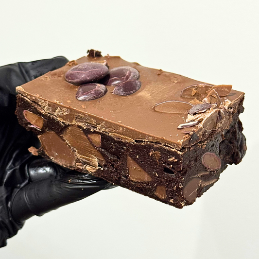 A hand wearing a black glove holds a square slice of chocolate fudge with embedded chocolate chips and nuts against a plain light background.