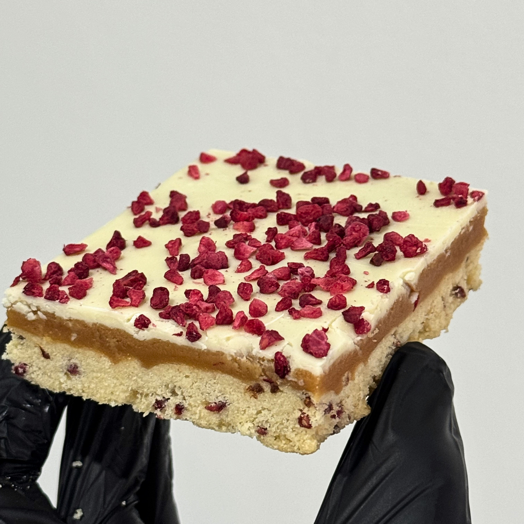 A square slice of layered dessert held up by black-gloved hands against a plain light gray background. The dessert has a pale cream top layer sprinkled with red dried fruit pieces, a caramel-like orange-brown middle layer, and a light beige crumb base.