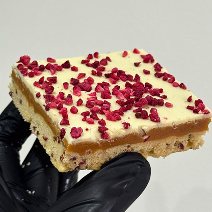 A square slice of layered dessert bar held by a hand wearing a black glove. The dessert has a light beige crumb base, a glossy amber caramel layer, and a thick off-white cream top generously sprinkled with red dried fruit pieces.