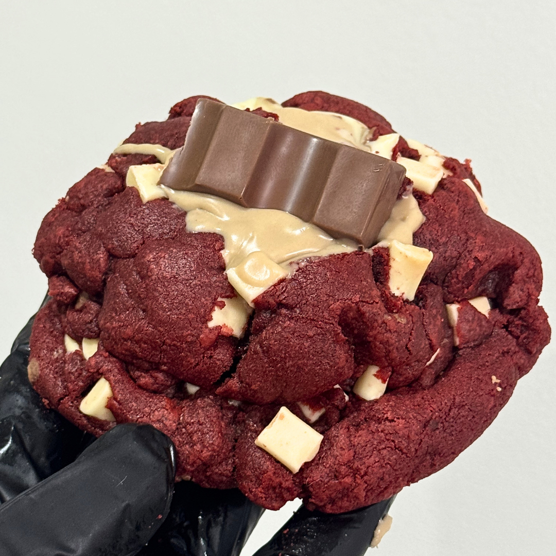 A close-up of a red velvet chocolate chip cookie topped with a chocolate bar and cream-colored drizzle, held by a black gloved hand against a plain light background.