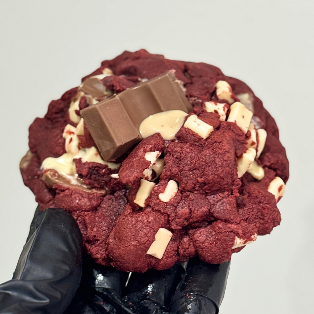 A close-up of a large red velvet chocolate cookie topped with a milk chocolate bar and white chocolate pieces, held by a hand wearing a black glove against a plain light gray background.
