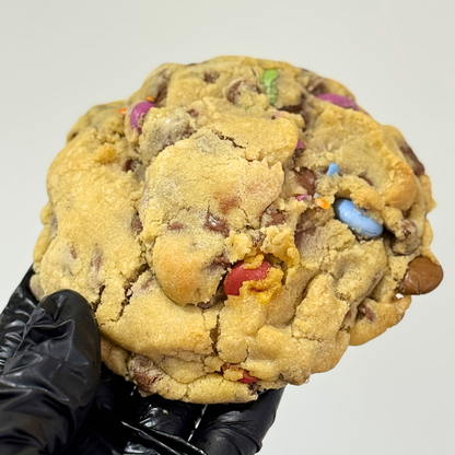 A large chocolate chip cookie with colorful candy pieces, held by a hand wearing a black glove against a plain light gray background.