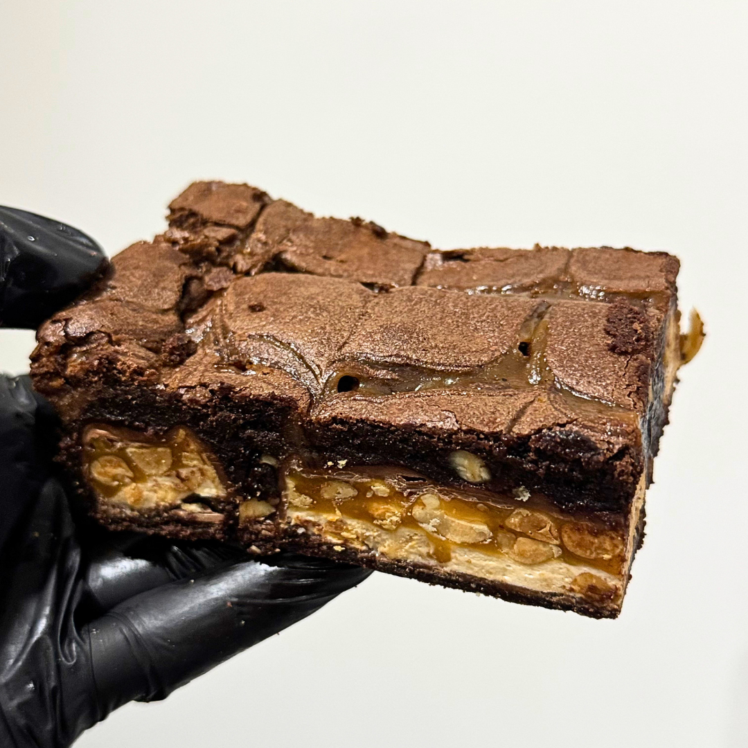 A close-up of a hand wearing a black glove holding a square slice of chocolate brownie with caramel and nuts, against a plain light background.