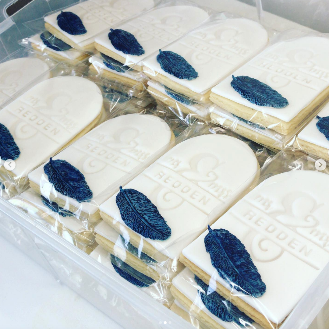 A clear plastic tray holding multiple individually wrapped rectangular cookies decorated with white icing, embossed text, and a dark blue feather motif.