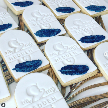 A close-up of several rectangular sugar cookies with white icing, embossed with a heart and the name 'HEDDEN,' decorated with dark blue feather stamps, arranged on a gold-toned tray.