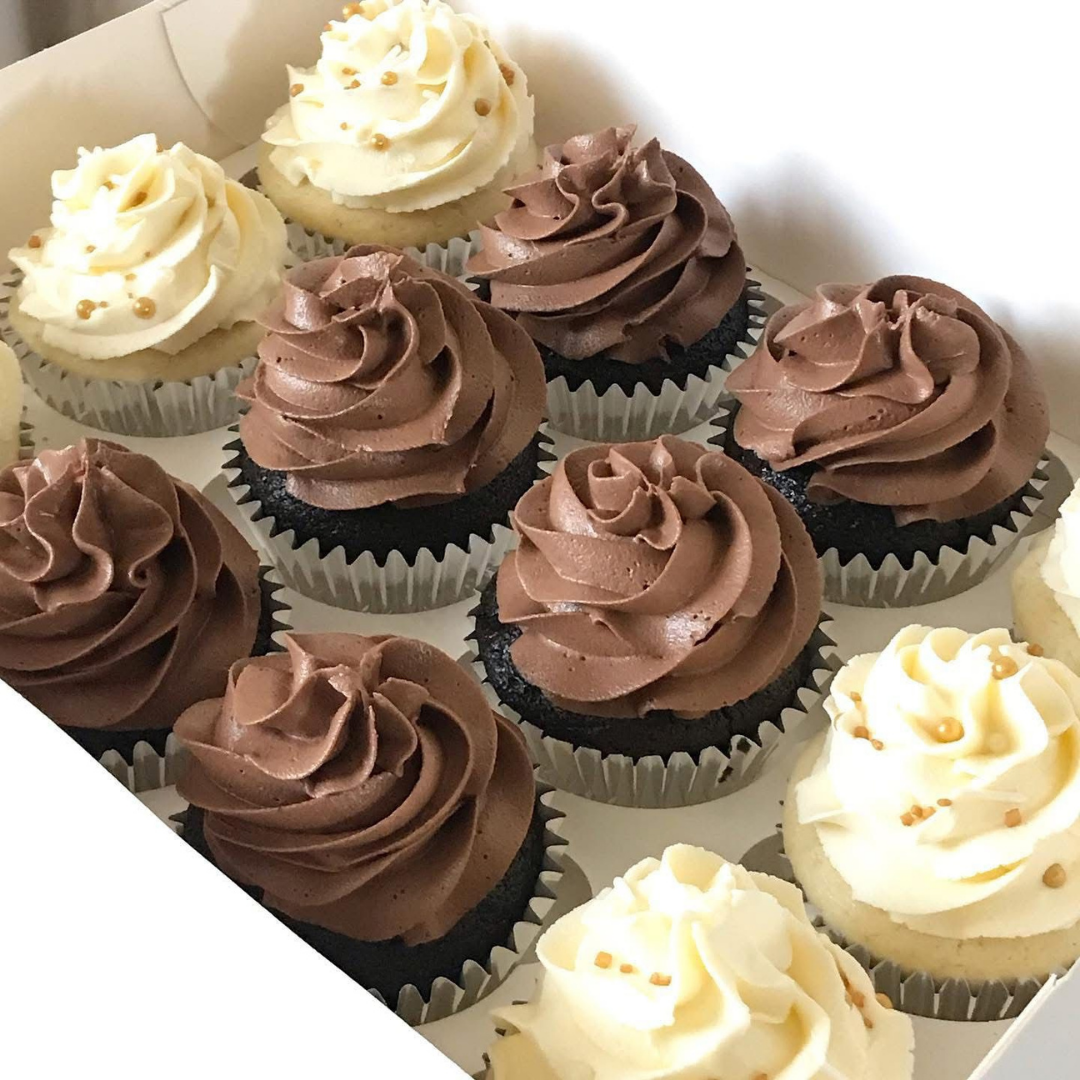 An assortment of frosted cupcakes in a white bakery box, featuring swirls of chocolate brown and creamy beige frosting with small sprinkle details.