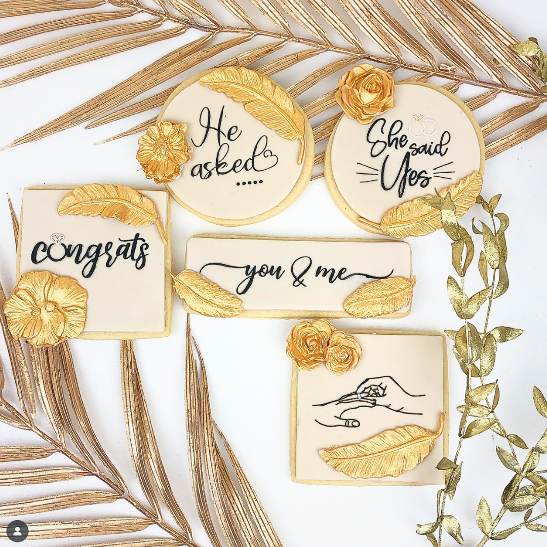 A flat-lay of five personalized sugar cookies decorated with gold leaf accents and black script text, styled with gold palm leaves on a white background.