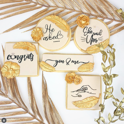 A flat-lay of five personalized sugar cookies decorated with gold leaf accents and black script text, styled with gold palm leaves on a white background.